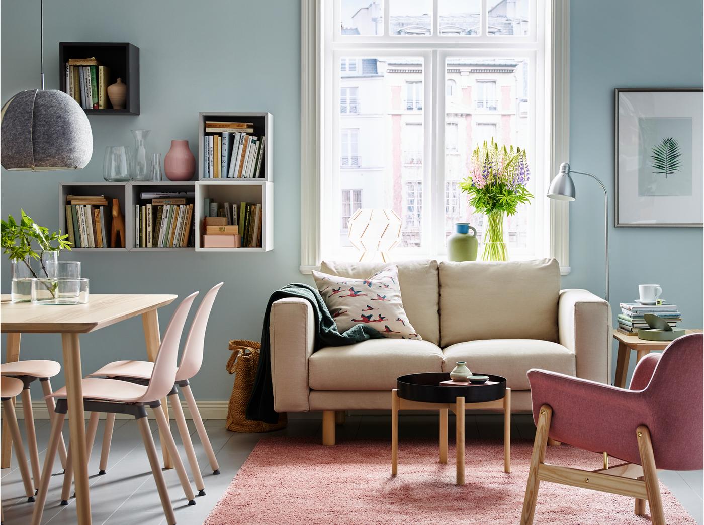 مساحة مفعمة بالدفء في غرفة جلوس / مساحة طعام صغيرة ذات تصميم مفتوح تتضمن كنبة بمقعدين NORSBORG بلون بيج Edum وكرسي بذراعين VEDBO بلون بني_وردي Gunnared بجوار طاولة قهوة YPPERLIG بلون رمادي داكن على خلفية زرقاء.