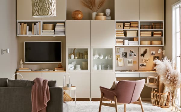 Mueble color crema BESTÅ, donde encuentras un televisor, libros y decoración. Está en un living donde hay una silla rosa o rosada con soporte de madera, un sofá gris oscuro con una frazada rosada encima y una alfombra gris clara.