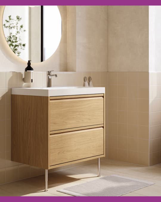 Mueble de baño en madera clara con lavamanos blanco, grifería metálica y espejo redondo sobre pared de cerámicos beige.
