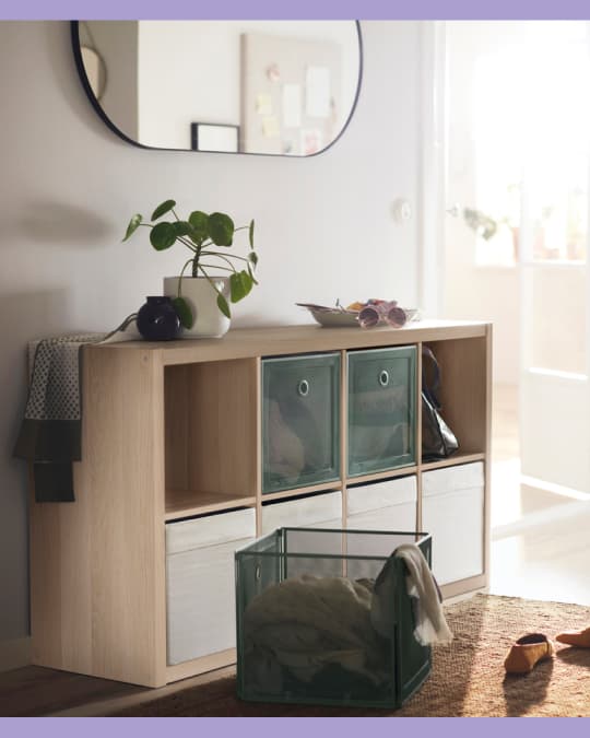 Mueble modular de madera clara con varios cubos, utilizado para almacenamiento con canastos, libros y objetos decorativos; sobre el mueble hay plantas y cerámicas.