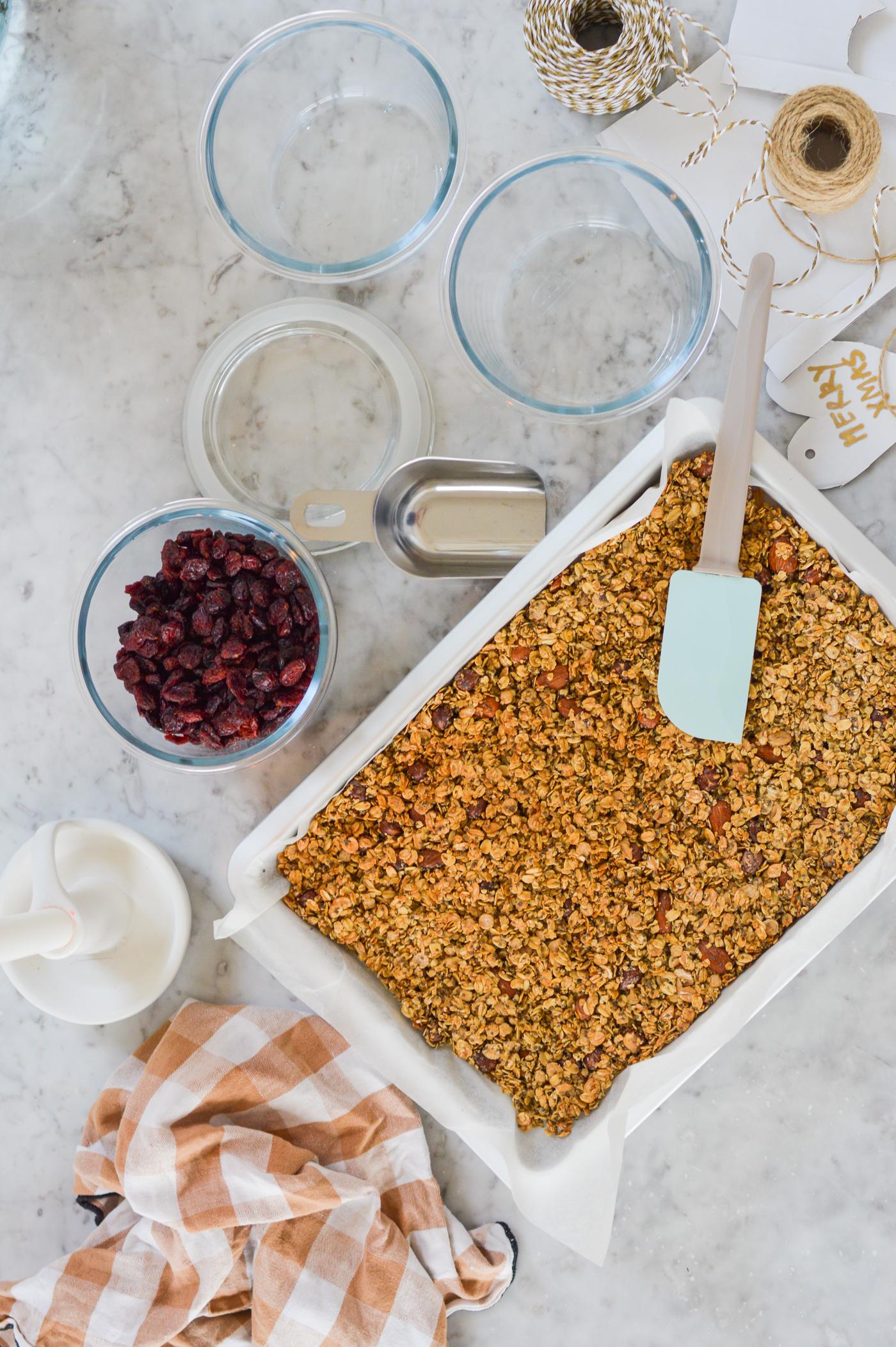 Muesli dans un plat à gratin avec une spatule bleue sur un plan de travail en marbre. Entouré de bols contenant des canneberges séchées, de bocaux et d'un torchon rayé, ce qui crée une ambiance chaleureuse dans la cuisine.