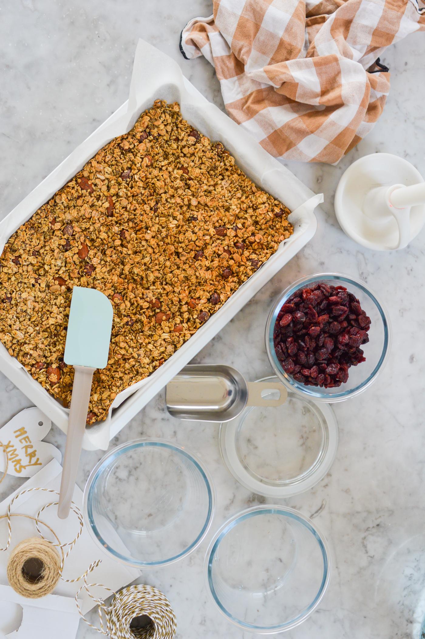 Muesli in a baking dish with a blue spatula on a marble worktop. Surrounded by bowls of dried cranberries, jars and a striped cloth, evoking a cosy kitchen scene.