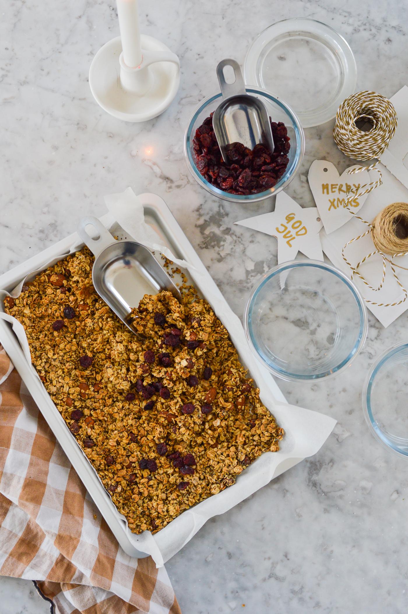 Muesli in a white baking dish on a marble surface, surrounded by a burning candle, a jar of cranberries, string, Christmas decorations and a kitchen towel.