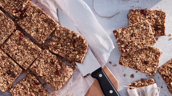 Muesli tyčinky na bílém, pečicím papíru a dřevěném prkénku, vedle kuchyňského nože a dvou zabalených plátků.