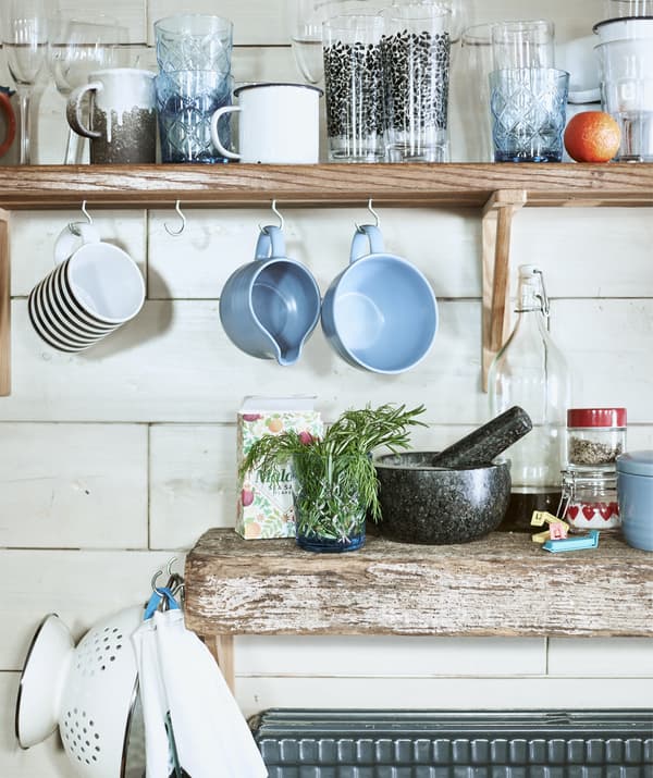 Mugs and cups on a shelf and hanging from hooks above a wooden worktop.