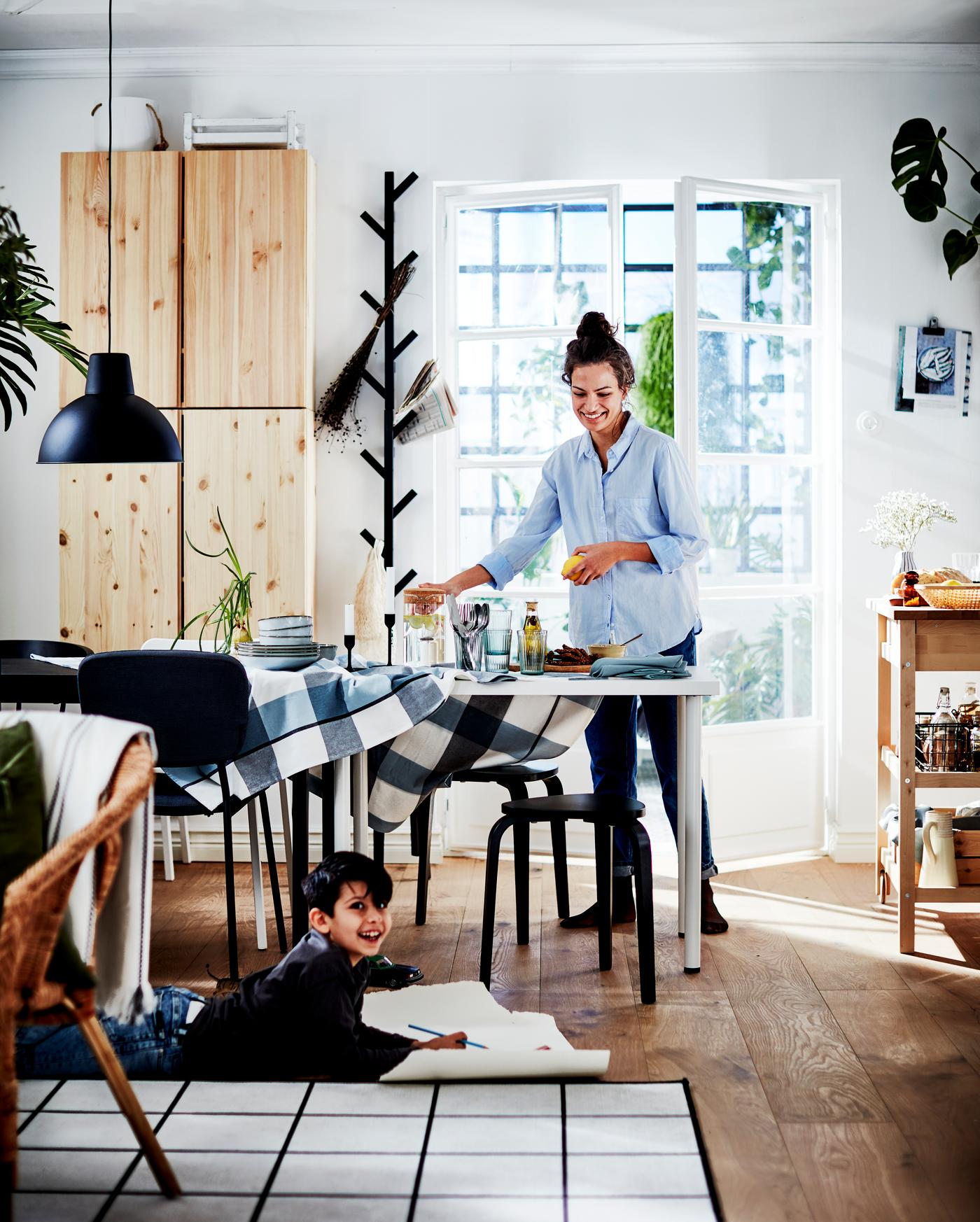 Mujer con camisa celeste ordenando la mesa del comedor mientras mira a su hijo en el suelo sobre una alfombra y dibujando en un papel.