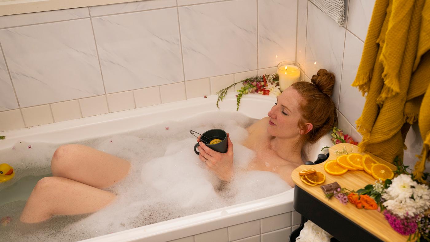 Mujer dándose un baño relajante en una bañera con espuma y con velas y plantas aromáticas alrededor.