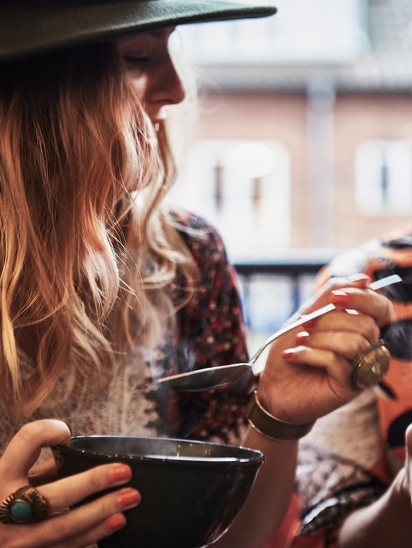 Mujer pelirroja comiendo comida caliente de un cuenco con una cuchara. Lleva un sombrero y joyas.