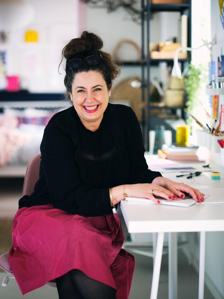 Mujer riéndose sentada en un escritorio blanco con sus manos sobre un cuaderno. Viste con un chaleco negro y una falda fucsia.