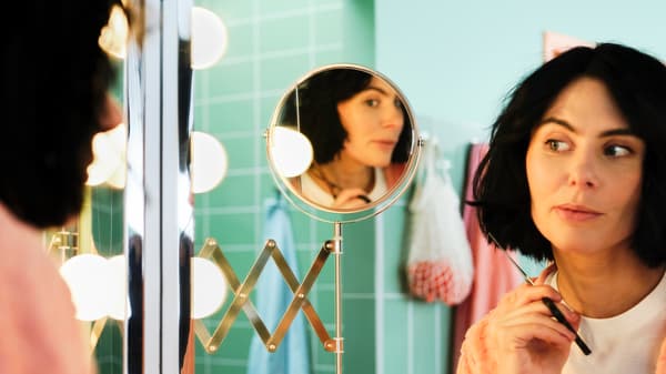 Mulher a segurar numa tesoura, a olhar para um espelho regulável FRÄCK enquanto corta o próprio cabelo numa casa de banho verde.