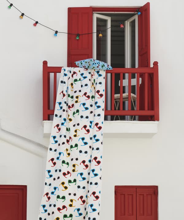Multicolour fabric with sunglasses print hanging from a red balcony on a white wall, with multicolour string lights.