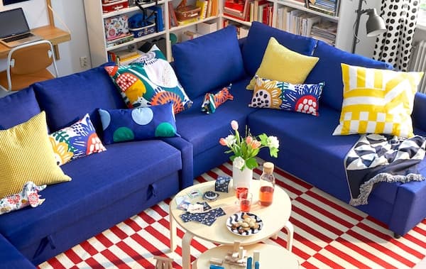 Multifunkčná modrá rohová pohovka IKEA FRIHETEN s farebnými vankúšmi, dva okrúhle konferenčné stolíky a pásikavý koberec.