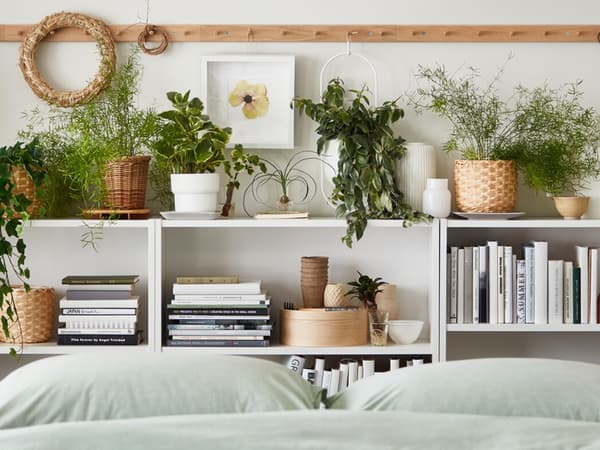 Multiple potted plants, real and artificial, stand amid books and decoration on shelves by a bed with BERGPALM bed linen.