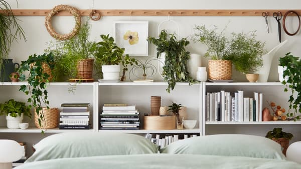 Multiple potted plants, real and artificial, stand amid books and decoration on shelves by a bed with BERGPALM bed linen.