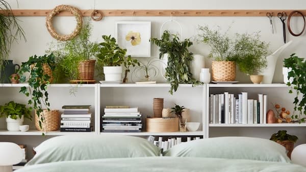 Multiple potted plants, real and artificial, stand amid books and decoration on shelves by a bed with BERGPALM bed linen.