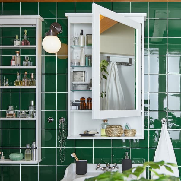 Mur de carreaux verts avec armoire à miroir HEMNES blanche fixée au mur ; la porte est ouverte pour laisser entrevoir des produits sur les tablettes à l’intérieur.