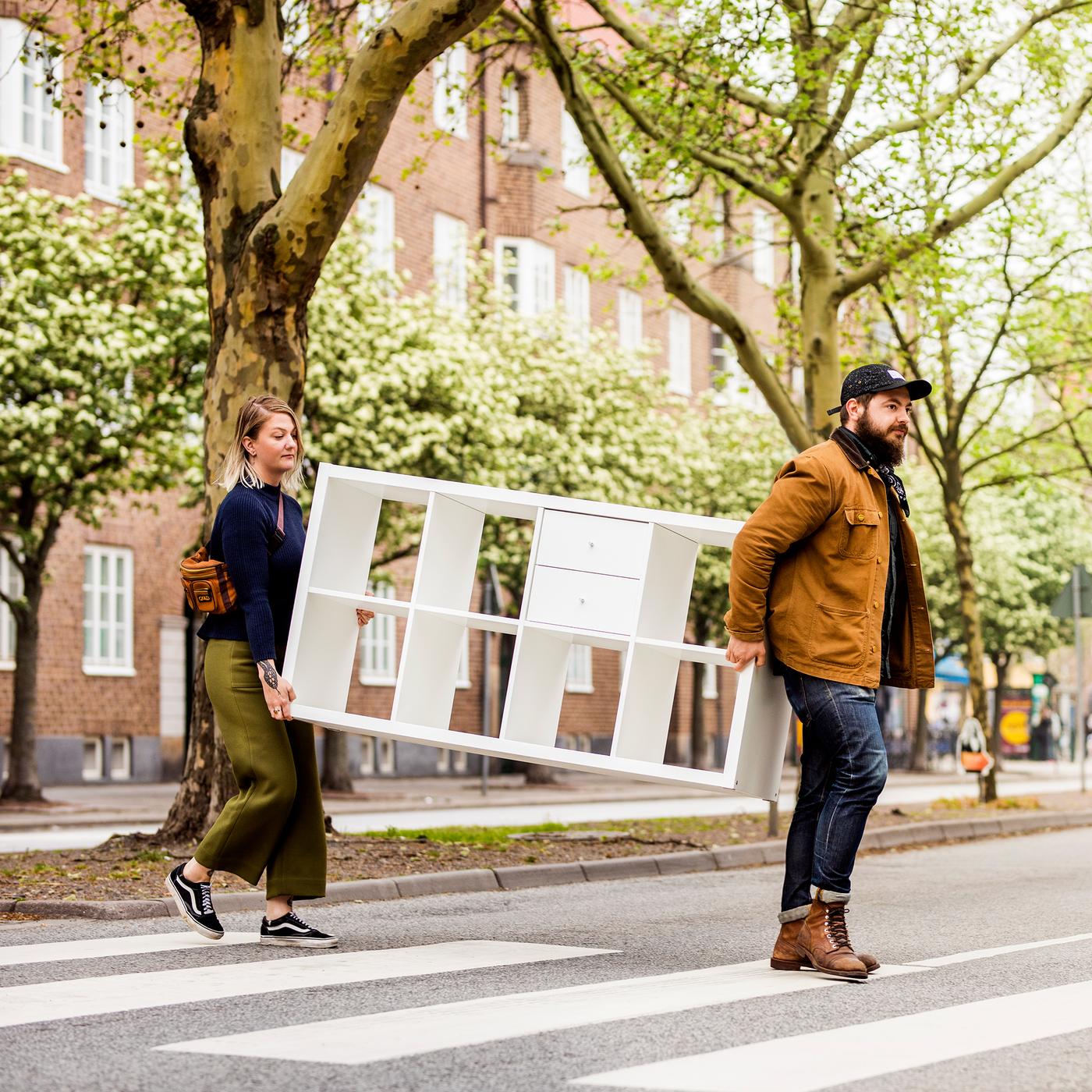 Muškarac i žena prelaze ulicu i nose KALLAX policu.