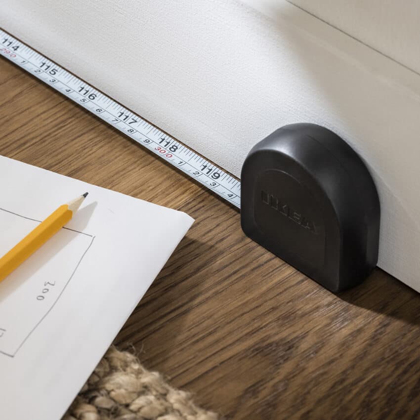 Musta mittanauha tummalla puisella työtasolla. Mitan vieressä näkyy suunnitelman nurkka sekä keltainen lyijykynä.