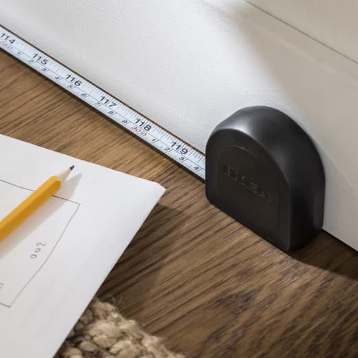 Musta mittanauha tummalla puisella työtasolla. Mitan vieressä näkyy suunnitelman nurkka sekä keltainen lyijykynä.