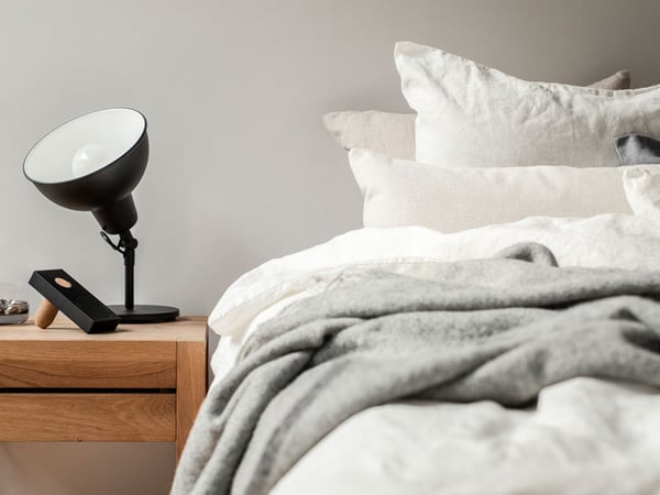 Musta valaisin yöpöydällä, sängyssä harmaat ja beiget vuodevaatteet.