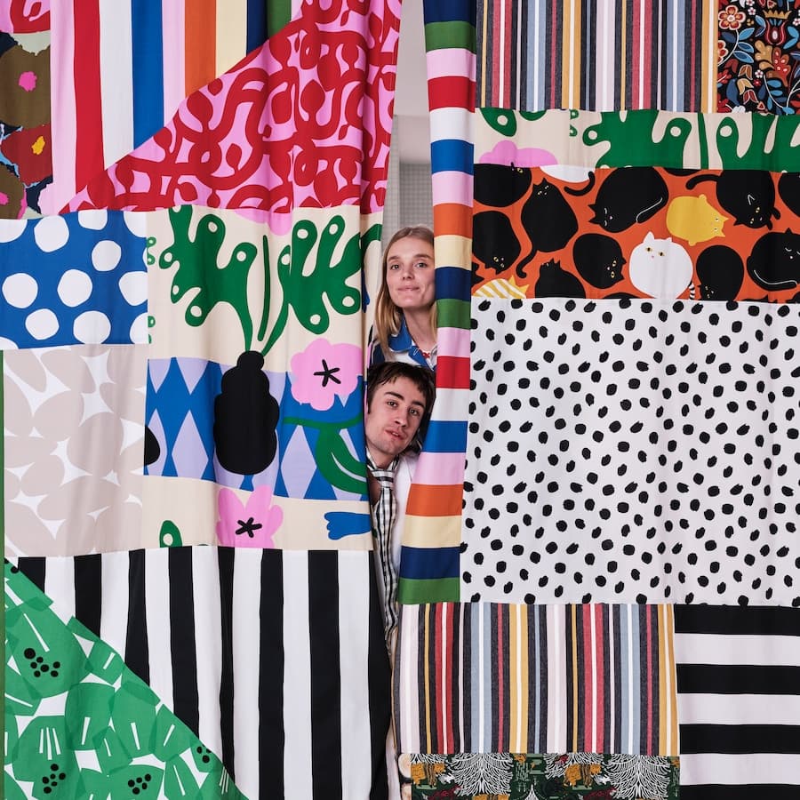 Muž a žena vykoukající zpoza závěsú sešitých z mnohých různých kusú divoce barevných a vzorovaných látek.