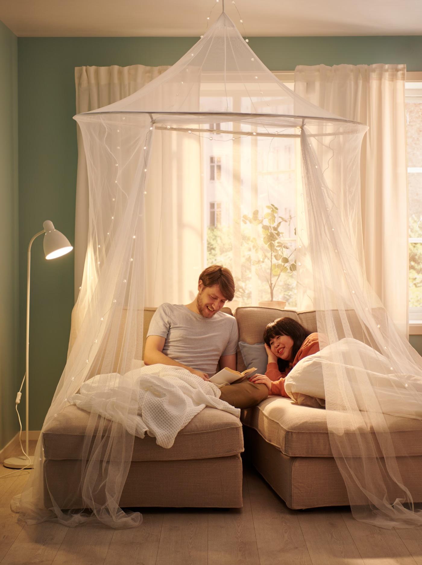 Muž čtoucí ženě příběh z knihy na pohovce pod síťovanou moskytiérou.
