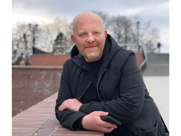 Muž s blond bradou v tmavo šedej bunde, opierajúci sa o múr vonku.