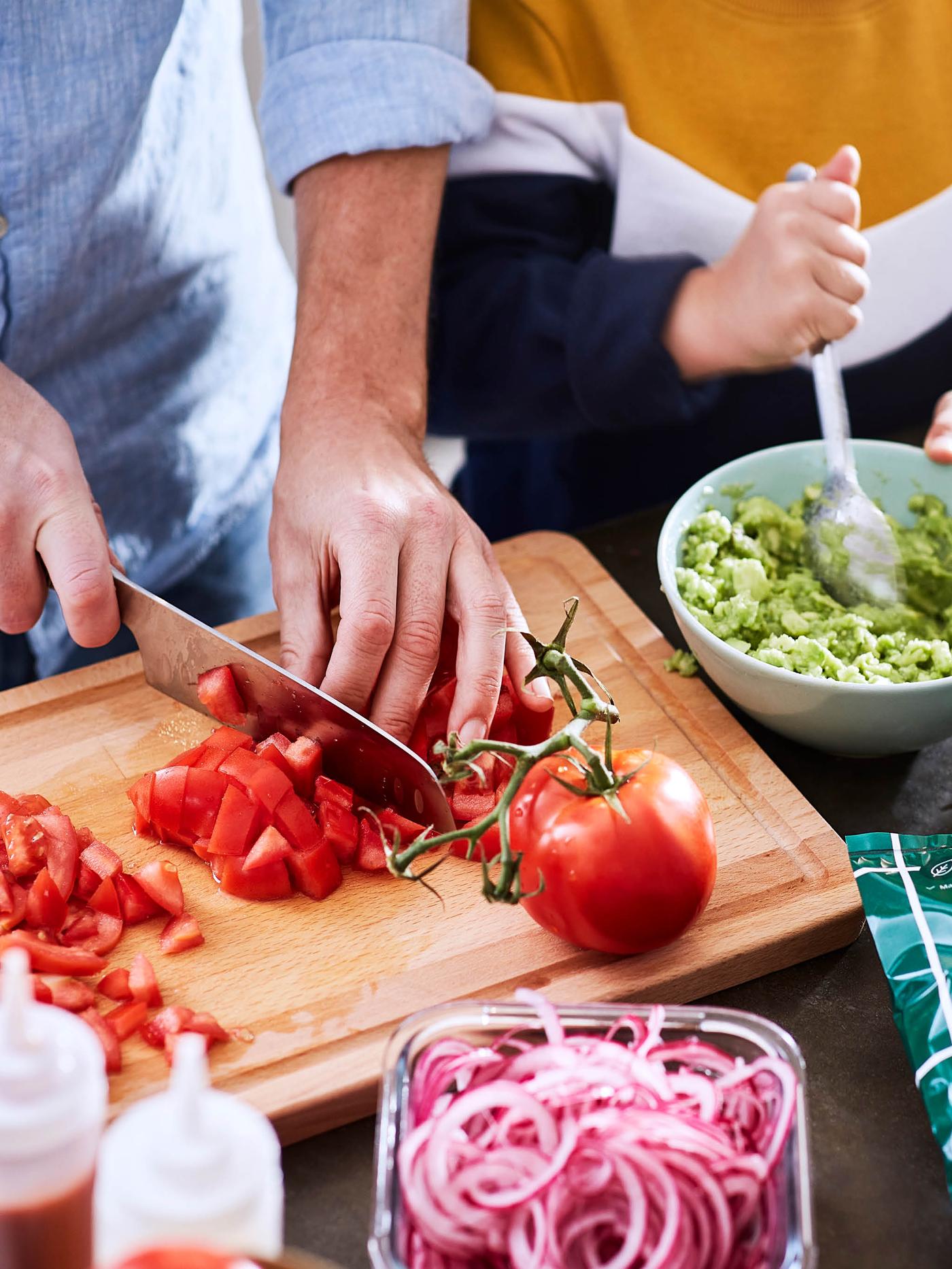 Muž v modrej košeli krája na doske APTITLIG paradajky, vedľa je chlapec, ktorý roztláča v miske lyžicou avokádo na guacamole.