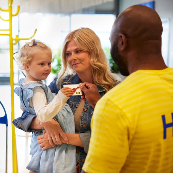 Muž v zaměstnanecké uniformě IKEA předává dítěti, které drží jeho matka, refundační kartu.