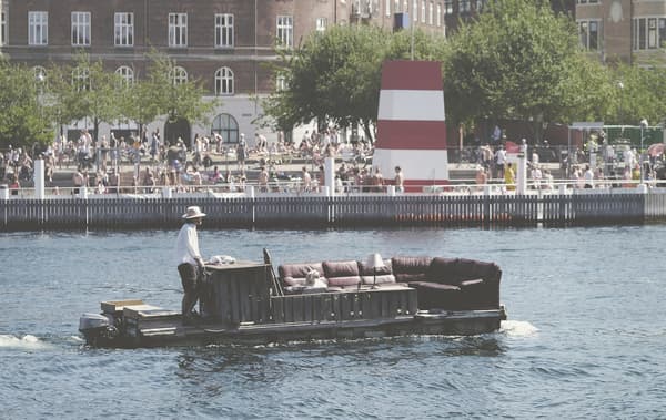 Muž veze velkou pohovku na raftu, kolem davy lidí a na břehu stromy a budovy