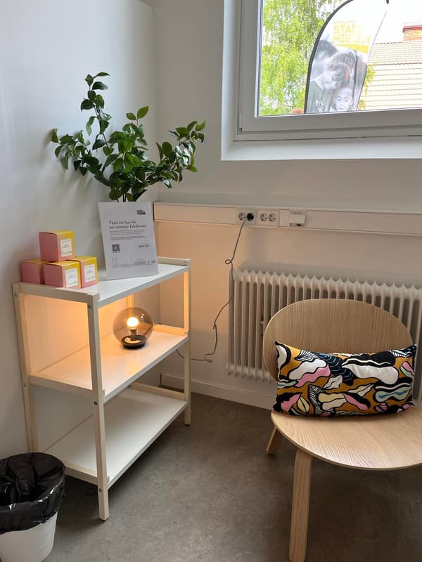 Mysig hörna med vit hylla, växt, tända ljus, färgglad kudde på bord och fönster med utsikt mot grönska i Stadsmissionen i Västerås.