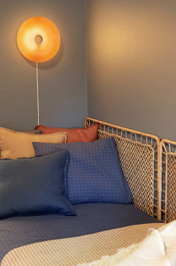 Mysigt hörn med dagbädd, färgglada kuddar och rottinggavel mot blå vägg. En rund vägglampa med varm orange belysning skapar en inbjudande stämning.