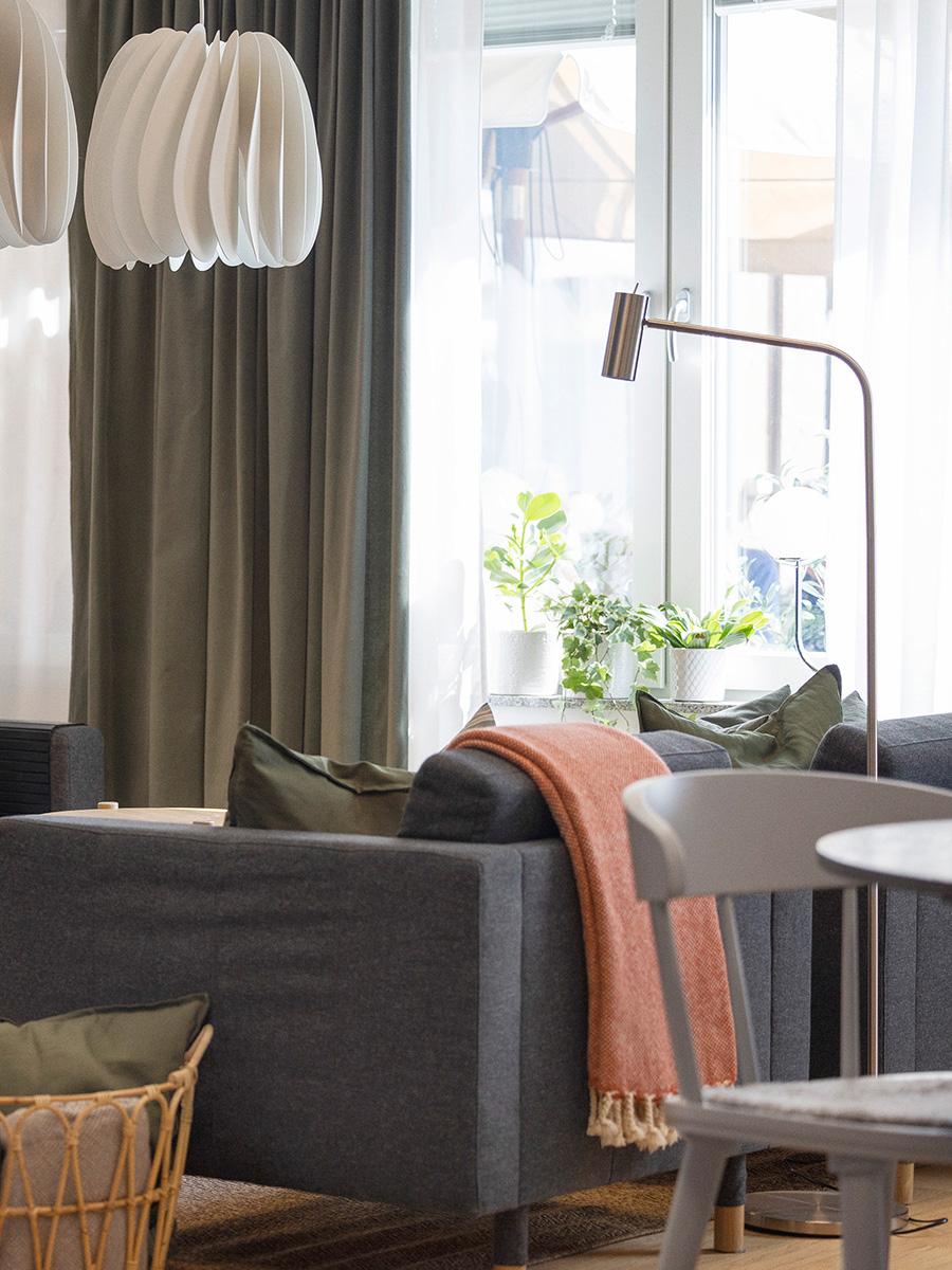 Mysigt vardagsrum med mörkgrå soffa, gröna kuddar och orange filt. Skulptural taklampa, småbord, grå stol och stora fönster med gardiner. Gröna växter vid fönstret.