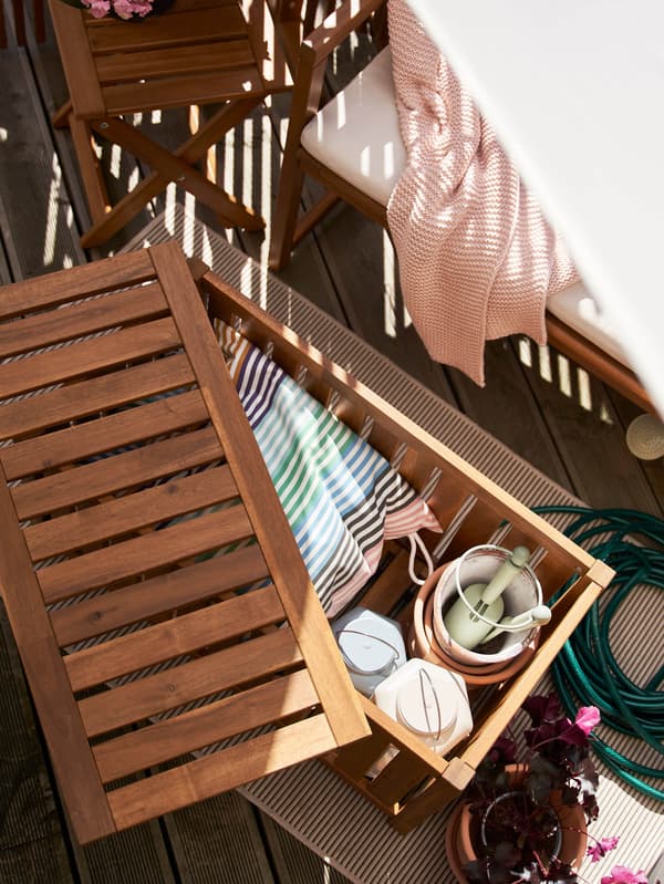 Na balkóne stojí pootvorený úložný diel NÄMMARÖ, vnútri vidno vankúš, kvetináče, záhradnícke náčinie a dva lampáše.