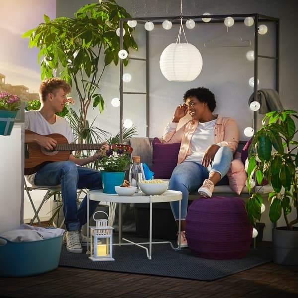 Na balkonie ławki z pergolą SVANÖ wiszą zasilane energią słoneczną lampy SOLVINDEN. Ławka jest udekorowana girlandą. Dziewczyna siedzi na ławce a chłopak gra na gitarze.