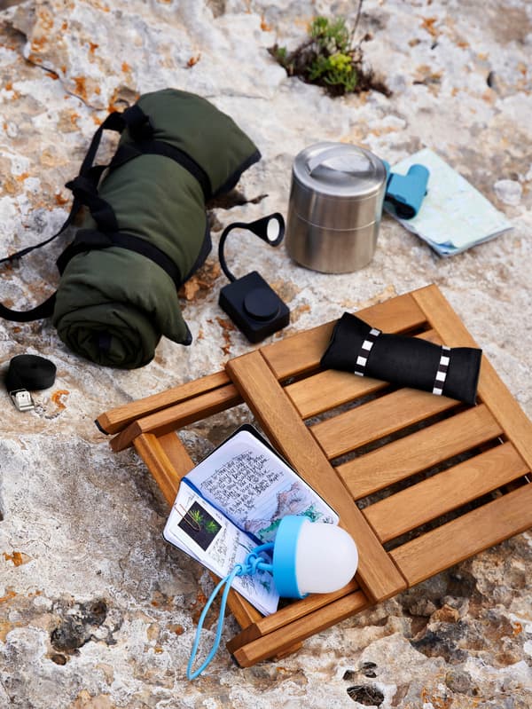 Na bielej skale je skladacia stolička NÄMMARÖ, dvojposchodová termoska FÖRSKAFFA, LED osvetlenie SOMMARLÅNKE, cestovný príbor FINSKUREN a pikniková deka FJÄLLMOTT.