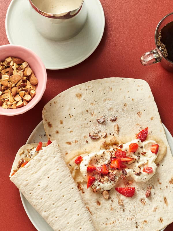 Na červeném stole leží bílý talíř leží s dvěma plátky tenkého chleba GRÄDDAT s vanilkovou omáčkou KAFFEREP, jahodami, zakysanou smetanou a oříšky, vedle stojí miska oříšků, konvička vanilkové omáčky a hrnek černé kávy.