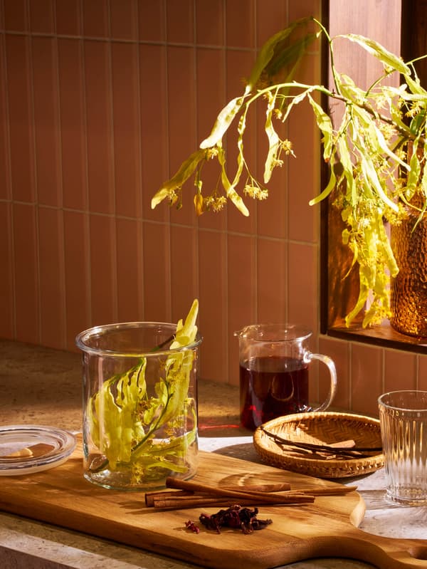 Na doske na krájanie ARTISTISK stojí dóza IKEA 365+ s lipovým kvetom, vedľa toho sú položené tri kúsky škorice a kôpka badiánu, v pozadí je odmerka VARDAGEN plná čaju a pohár VARDAGEN.