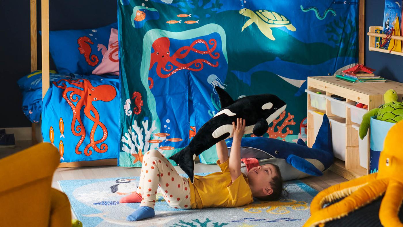 Na koberci BLÅVINGAD leží chlapec a dvíha nad seba plyšovú kosatku BLÅVINGAD, za ním na koberci leží plyšová veľryba BLÅVINGAD, v pozadí je posteľ s modrým povlečením BLÅVINGAD a baldachýnom KURA.
