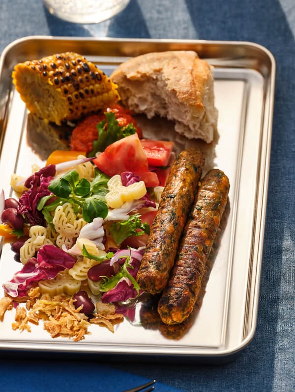 Na modrém ubruse leží kovový tácek a dvěma zeleninovými hot dogy KORVMOJ vedle porce těstovinového salátu, kousku chleba a grilované kukuřice.