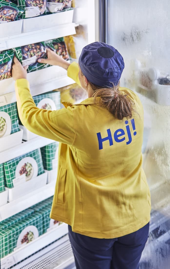 Na obrázku je zaměstnanec (prodavač/ka) v supermarketu, který doplňuje zboží do regálů. Má na sobě žlutou bundu s nápisem "Hej!" na zádech a tmavě modrou čepici. Doplňuje pravděpodobně mražené potraviny, které jsou v zelenobílých obalech s obrázky jídel na talíři. V pozadí je vidět zamrzlá stěna chladicího pultu.
