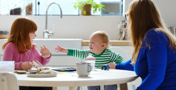 Na obrázku je žena se dvěma dětmi u kulatého stolu, pravděpodobně v kuchyni nebo jídelně. Starší dítě sedí na obyčejné židli, zatímco batole sedí v dětské vysoké židličce a natahuje ruce. Všichni se usmívají. Na stole je hrnek, láhev s pitím a malý talíř se svačinou.