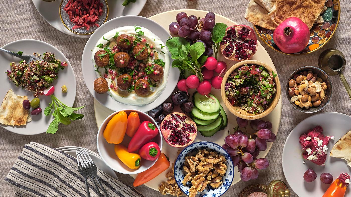 Na otočnom podnose SNUDDA je tanier s bezmäsitými guľôčkami HUVUDROLL a so zemiakovou kašou, miska s paprikami, zväzok reďkoviek, hrozno, granátové jablko, miska s orieškami a vedľa toho sú ďalšie taniere s jedlom.