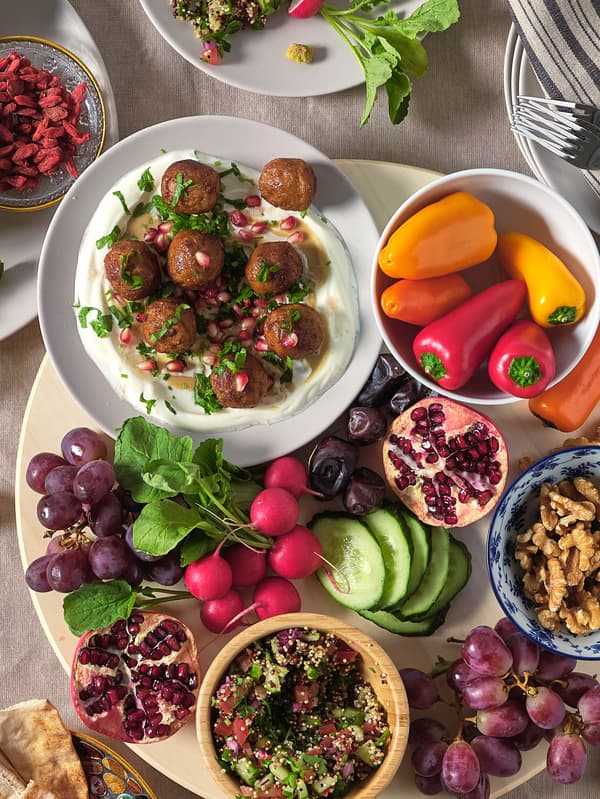 Na otočnom podnose SNUDDA je tanier s bezmäsitými guľôčkami HUVUDROLL a so zemiakovou kašou, miska s paprikami, zväzok reďkoviek, hrozno, granátové jablko a miska s orieškami.