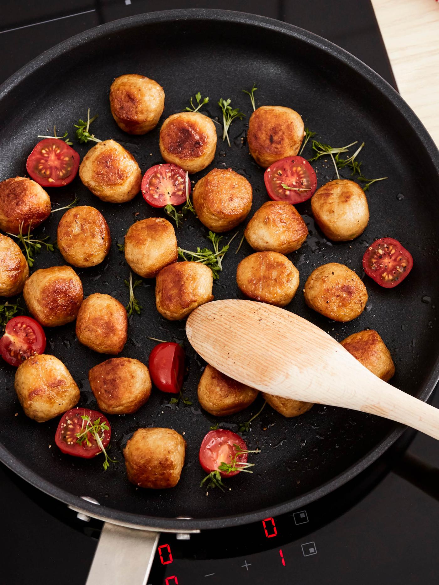 Na pánvi HEMKOMST míchá lžíce RÖRT kuřecí kuličky, půlky cherry rajčat a snítky čerstvé řeřichy.