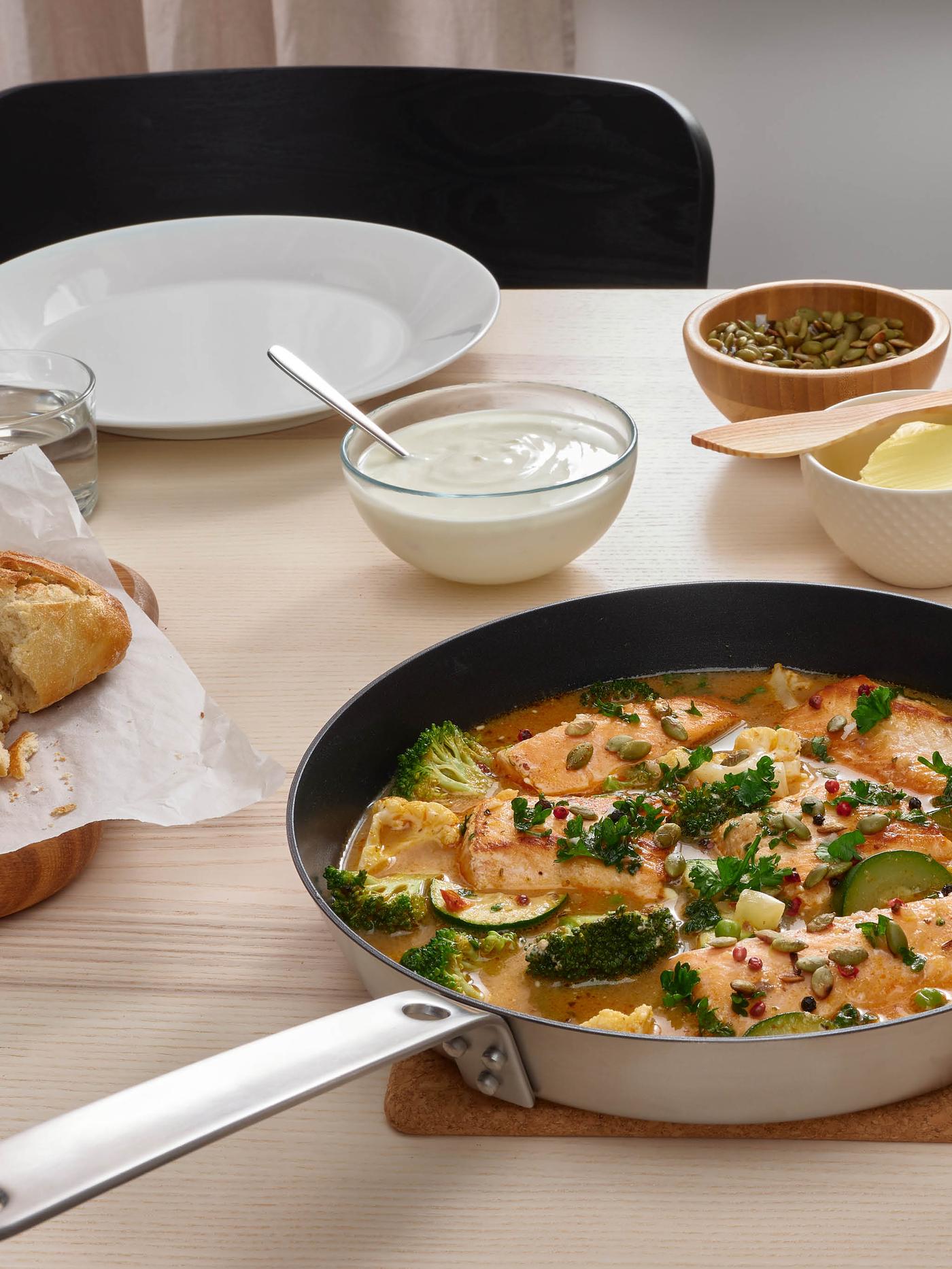 Na podložke pod hrnce HEAT je panvica HEMKOMST s lososom SJÖRAPPORT v kokosovej omáčke, v pozadí je biely tanier, pohár vody, miska s tekvicovými semienkami a miska s jogurtom, vedľa toho je na doske pečivo.