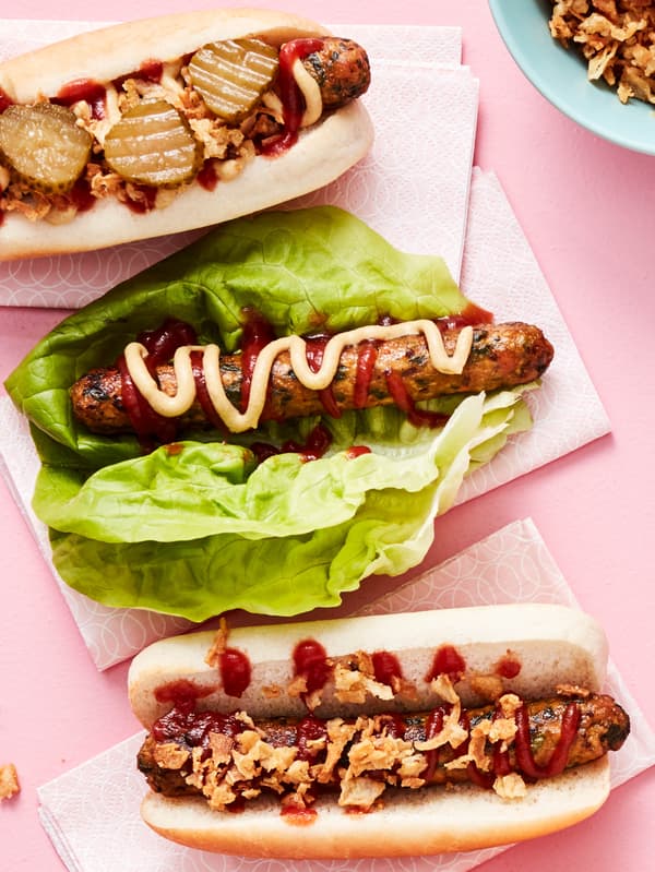 Na růžové ploše leží na bílých ubrouscích 3 obložené zeleninové hot dogy KORVMOJ, dva v housce, jeden v listu salátu.