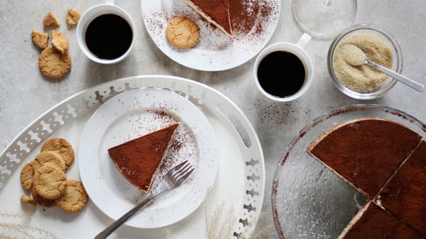Na servírovacím stojanu ARV BRÖLLOP je cheescake posypaný kakaem, vedle leží dva dezertní talíře OFANTLIGT s jednotlivými porcemi, dva hrnky s kávou a cukřenka s třtinovám cukrem.