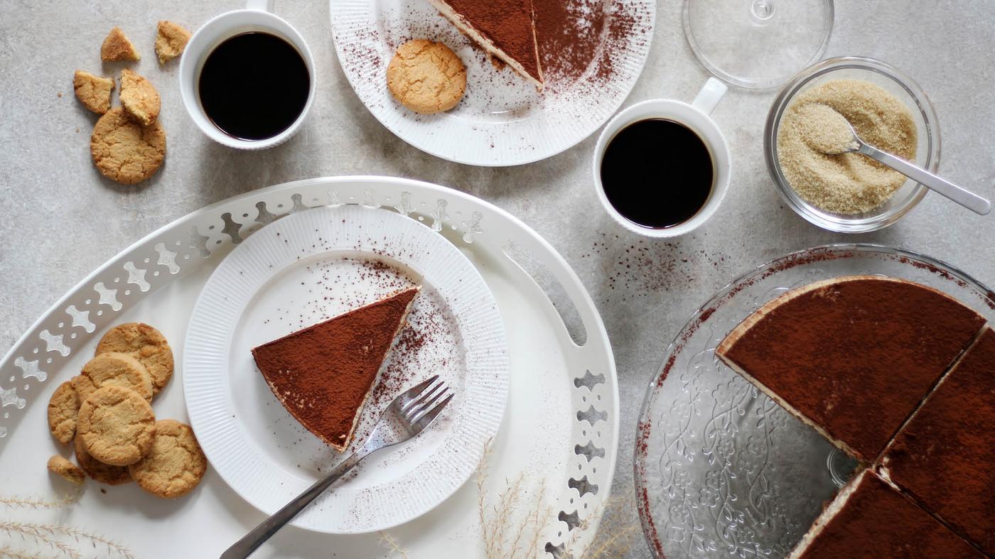 Na servírovacom stojane ARV BRÖLLOP je cheescake posypaný kakaom, vedľa toho sú dva dezertné taniere OFANTLIGT s jednotlivými porciami, dve šálky s kávou a nádobka s trstinovým cukrom.