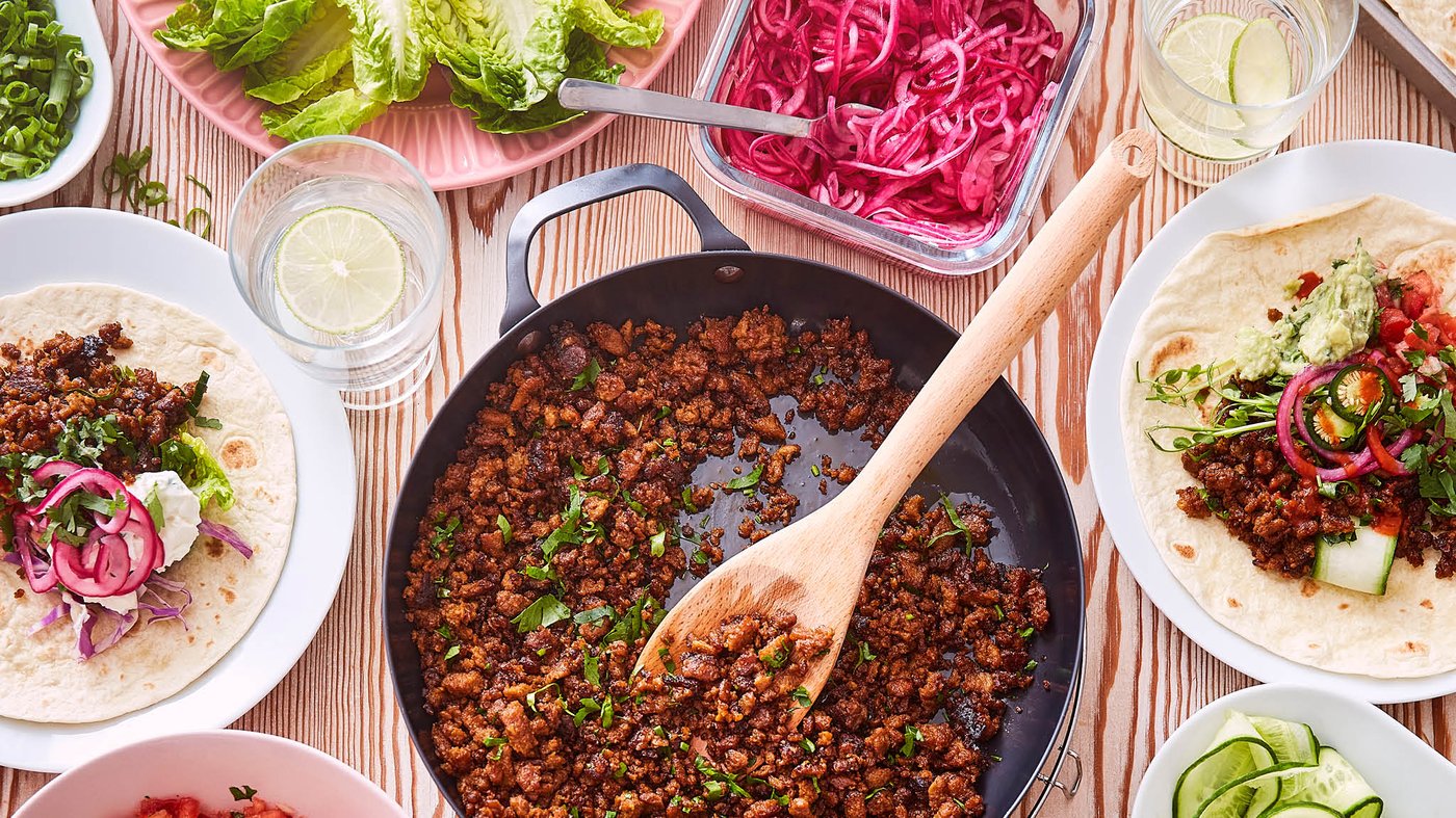 Na stole je panvica VARDAGEN plná bezmäsitej zmesi VÄRLDSKLOK na tacos, okolo sú taniere s tortillami a miska s nakladanou červenou cibuľou.